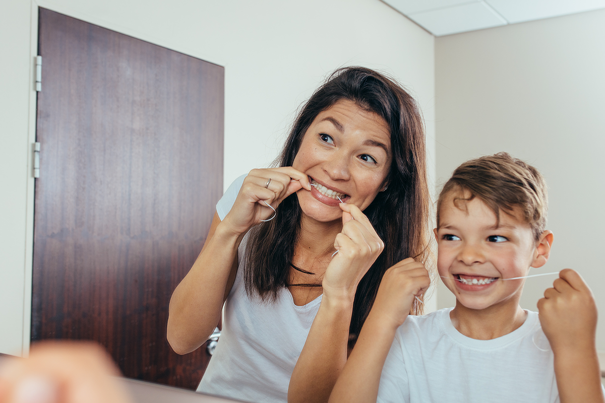 Mother and child brushing teeth together in bathroom, both smiling at camera.