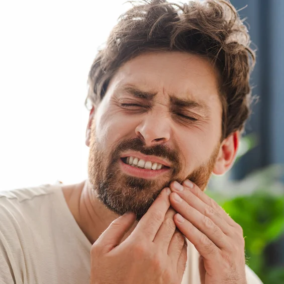 Man experiencing tooth pain, holding cheek with hand outdoors in daylight.