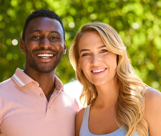 Two smiling people posing outdoors with green foliage background for dental care practice.
