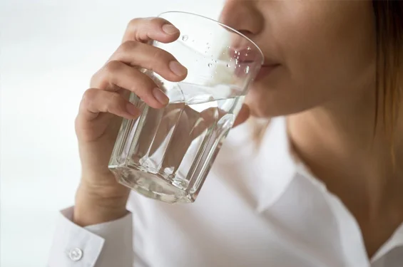 Person drinking water from a glass, wearing white shirt against light background.
